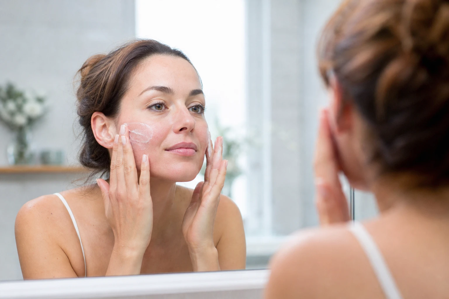 Femme appliquant une crème apaisante sur son visage marqué par la rosacée dans une salle de bain lumineuse